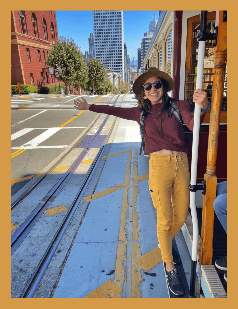 A woman stands on a cable car, smiling, wearing a hat, sunglasses, maroon top, and mustard pants, with city buildings in the background.